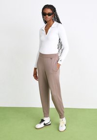 Polo blanc à manches longues avec un col, associé à un pantalon tapered marron clair et des lunettes de soleil noires. Des baskets noir et blanc complètent le look.