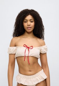 Σετ μπικίνι με τοπ έξω από τον ώμο σε κρεμ χρώμα με κόκκινες πουά, διαθέτει κόκκινη κορδέλα και κοντό, βολάν κάτω μέρος. Υφή υφάσματος.