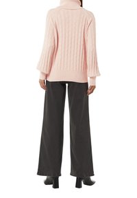 Rosa Kabelstrickpullover mit hohem Kragen, lockeren Ärmeln und geripptem Saum, kombiniert mit weiten, dunkelgrauen Hosen und schwarzen Absatzschuhen.