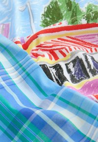 Tissu à carreaux bleu et vert superposé à un tissu coloré avec des motifs rouges, jaunes, noirs et arbres, montrant la texture et les plis.