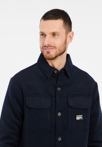 Tamsiai mėlynas švarkas, pagamintas iš minkštos medžiagos, su mygtukų užsegimu, dviem krūtinės kišenėmis ir logotipu ant kairės kišenės.