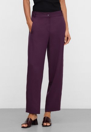 Persona che indossa pantaloni a gamba larga di colore viola scuro con tasche laterali e sandali aperti con tacco largo scuro, su uno sfondo semplice.