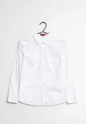 Witte blouse met lange mouwen, ruches bij de kraag en knoopsluiting, hangend aan een zwarte hanger tegen een witte achtergrond.