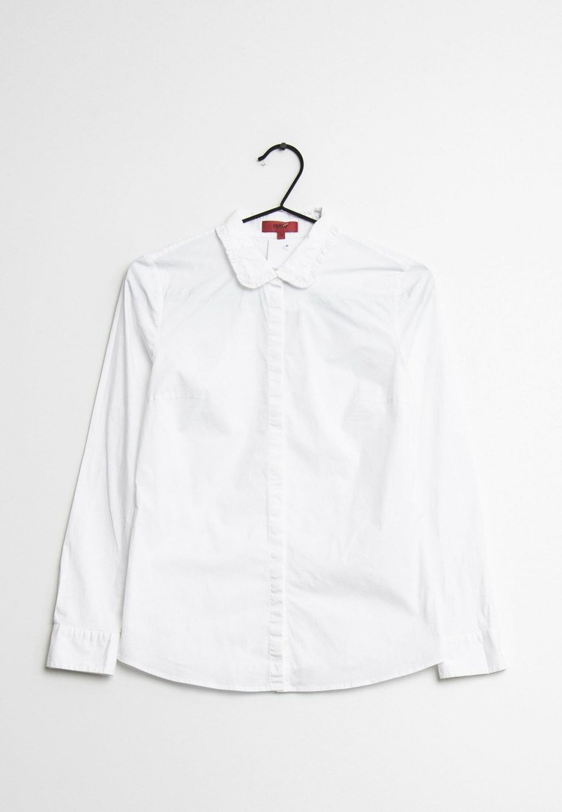 Blouse blanche à manches longues avec col à volants et patte de boutonnage, suspendue sur un cintre noir sur un fond blanc.