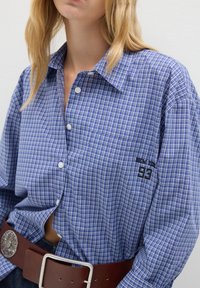 Donna che indossa una camicia a quadri blu con bottone, con ricamo "New York 93" sulla tasca del petto e una larga cintura di pelle marrone con una grande fibbia d'argento.