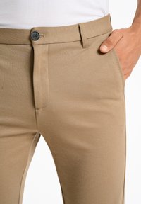Chinos beiges en tissu lisse et texturé, avec une fermeture à bouton et des poches latérales, mettant en valeur une coupe ajustée et des lignes épurées.