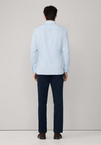 Lichtblauwe blouse met lange mouwen en knoopmanchetten, in combinatie met donkerblauwe broek en bruine schoenen, gezien vanaf de achterkant. Glad stoftextuur.