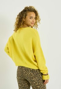 Mujer con cabello rizado que lleva un suéter amarillo brillante y pantalones con estampado de leopardo, mirando por encima del hombro con una ligera sonrisa.