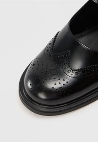 Scarpa di pelle nera con punta arrotondata, caratterizzata da perforazioni brogue e dettagli di cucitura. Texture liscia e suola piatta.