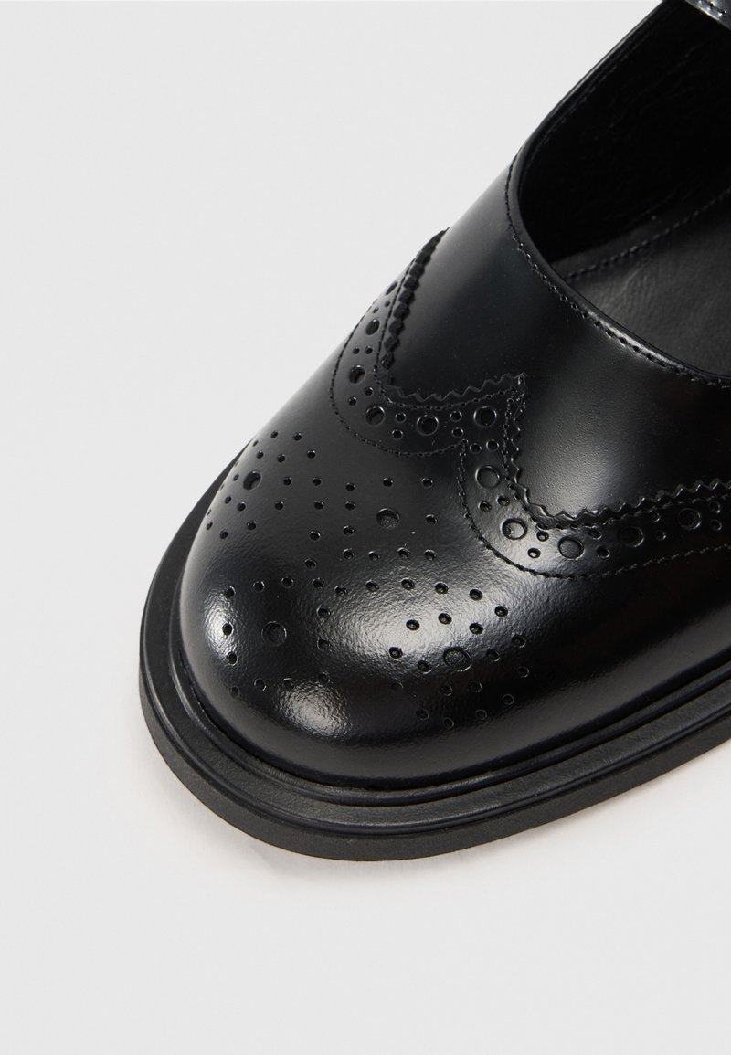 Chaussure en cuir noire avec un bout arrondi, présentant des perforations de brogue et des détails de couture. Texture lisse et semelle plate.