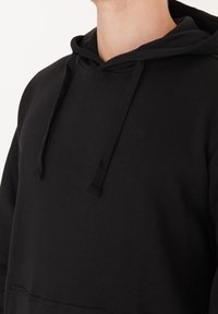 Lähikuva henkilöstä, joka on pukeutunut mustaan huppariin, jossa on narut ja pieni brodeerauslogo rinnassa.