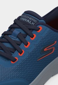 Κοντινό πλάνο μπλε αθλητικού παπουτσιού Skechers Go Walk με μαύρα κορδόνια, πορτοκαλί οφθαλμούς και πορτοκαλί λεπτομέρεια λογότυπου "S" πάνω σε υφασμάτινη επιφάνεια με υφή.