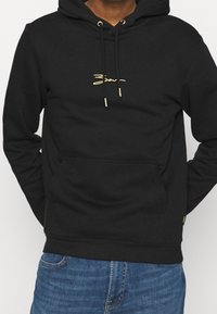 Sudadera con capucha negra hecha de una tela suave, presenta un bolsillo tipo canguro en la parte frontal, cordones dorados y un detalle de logo dorado minimalista.
