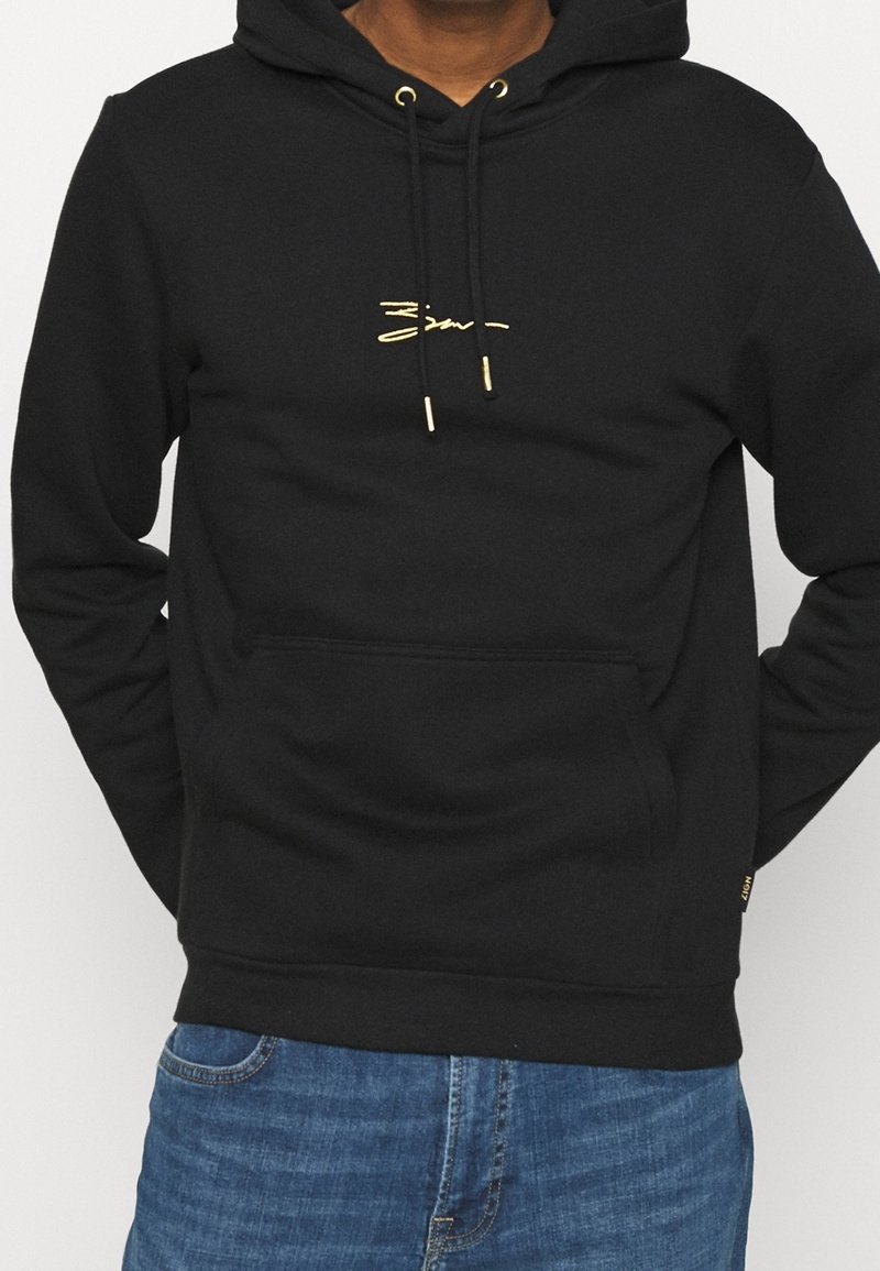 Sudadera con capucha negra hecha de una tela suave, presenta un bolsillo tipo canguro en la parte frontal, cordones dorados y un detalle de logo dorado minimalista.