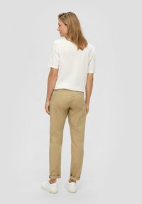 Losse witte katoenen T-shirt, beige chino's met opgerolde mouwen en witte sneakers, gezien van achteren tegen een grijze achtergrond.