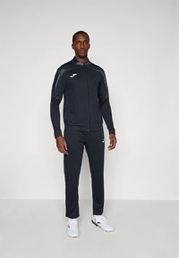 Tumši zils treniņtērps ar rāvējslēdzēju apakšdelma jaku un atbilstošām biksēm no sintētiskām materiāla. Uz krūtīm un bikšu sāniem redzams logotips.