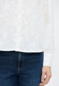 Λευκό πουκάμισο με κουμπιά και σχέδια από λουλουδένια δαντέλα, συνδυασμένο με σκούρο μπλε τζιν και εμφανές χέρι στη δεξιά πλευρά.