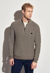 Pull en khaki avec un col zippé, texture c ôtelée et couleur unie. Présente un petit logo noir sur la poitrine. Assorti avec un jean foncé.