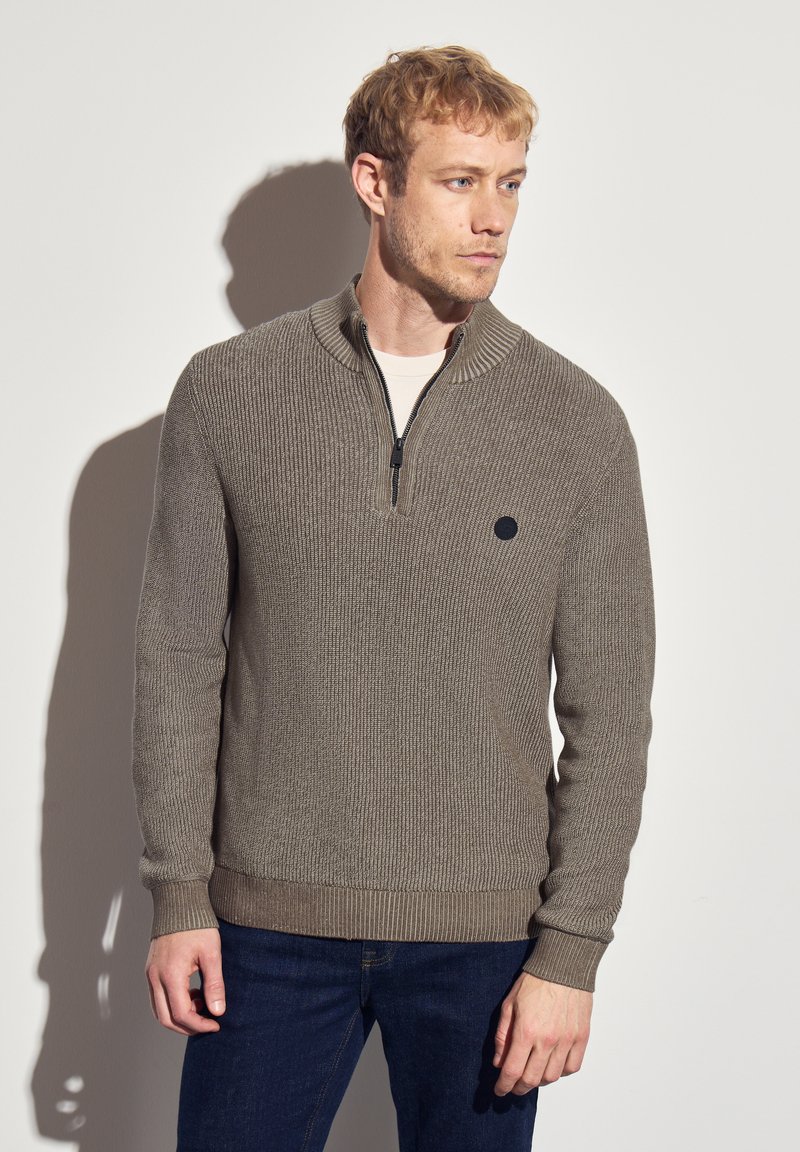 Pull en khaki avec un col zippé, texture côtelée et couleur unie. Présente un petit logo noir sur la poitrine. Assorti avec un jean foncé.