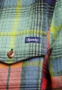 Camicia a quadri nei toni del verde, blu e arancione; realizzata in tessuto testurizzato con bottone marrone e etichetta ricamata "Superdry" sulla tasca.