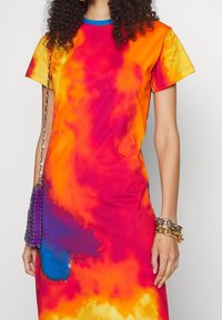 Barevné tričko s krátkým rukávem ve stylu tie-dye, které má oranžové, růžové a modré odstíny. Obsahuje modrý výstřih a stříbrné řetízkové detaily.