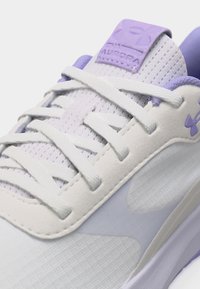 Λευκό και λεβαντέ αθλητικό παπούτσι με υφασμένες λευκές κορδόνια, ανθεκτικό ανώτερο μέρος από διχτυωτό υλικό και το λογότυπο Under Armour στη γλώσσα και στην πλευρά.