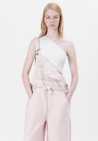 Rosa denimoveraller med én stropp, frontlomme og snor i midjen, satt sammen med en hvit topp med én skulder.