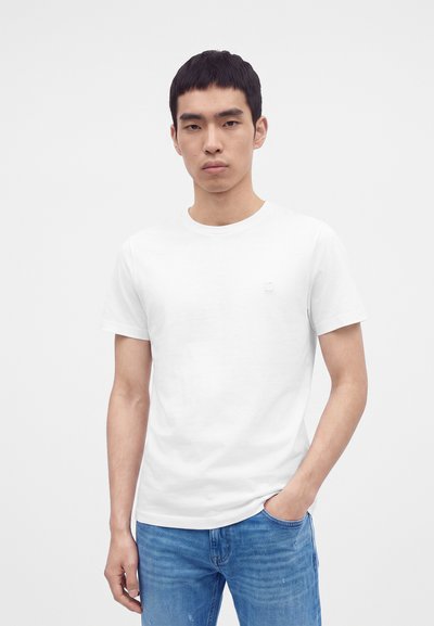 Jeune homme aux cheveux courts et foncés portant un t-shirt blanc uni et un jean bleu, debout devant un fond clair.