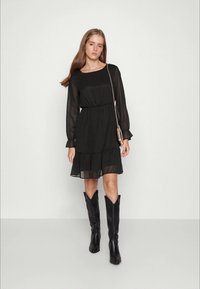 Vestido preto em chiffon com mangas longas, bainha com babados e cintura elástica. Combinado com botas pretas até ao joelho e uma pequena bolsa metálica a tiracolo.