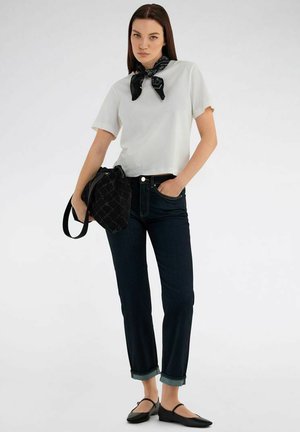 Frau steht, trägt ein weißes kurzärmliges Oberteil, schwarzen Halstuch, dunkle aufgekrempelte Jeans, schwarze Ballerinas und hält eine schwarze gemusterte Handtasche.