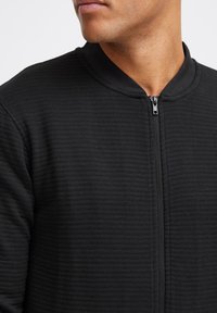 Chaqueta negra con cremallera de textura acanalada y cuello redondeado. La cremallera está ubicada en el centro, lo que contribuye a su diseño minimalista.