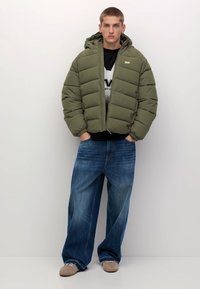 Ung man klädd i en olivgrön puffjacka, en grafisk svartvit skjorta, breda blå jeans och beige skor som står mot en slät vägg.