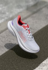 Chaussure de sport grise avec une tige respirante, des accents roses, une semelle rembourrée et un logo HOKA sur le côté. Design léger et moderne.