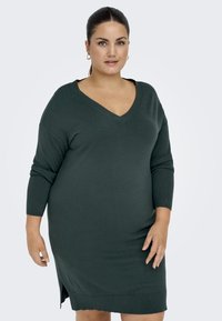 Robe tricotée vert foncé avec un décolleté en V, des manches 3/4, des fentes latérales et une coupe ajustée. Sa texture douce améliore le confort et le style.