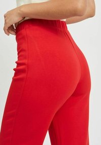 Rode, rekbare broek met een elastische tailleband, met een gladde textuur en een getailleerde pasvorm, die de achter- en zijaanzicht toont.