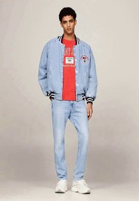 Heledasinine teksajakk mustade ja valgete triipudega, punane graafikaga T-särk, heledad teksad ja valged tossud. Seisev poos.