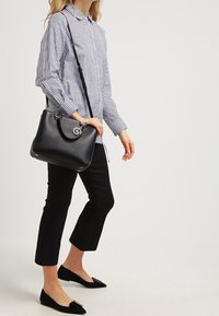 Borsa a mano in pelle nera con design curvo, hardware argentato e tracolla regolabile. Il modello indossa una camicia a righe blu e bianche e pantaloni neri.
