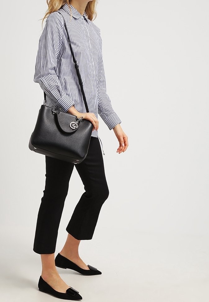 Borsa a mano in pelle nera con design curvo, hardware argentato e tracolla regolabile. Il modello indossa una camicia a righe blu e bianche e pantaloni neri.