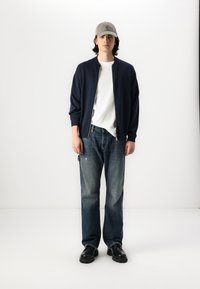 A jaqueta bomber azul-escura com fecho, por cima de uma t-shirt branca, combinada com jeans azuis desfiados e sapatos slip-on pretos. Boné cinza usado.