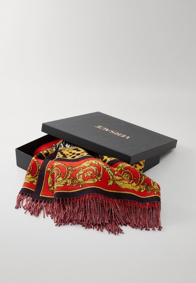Black box with gold "Versace" logo partially open, revealing a vibrant red, yellow, and black patterned scarf with fringed edges inside.