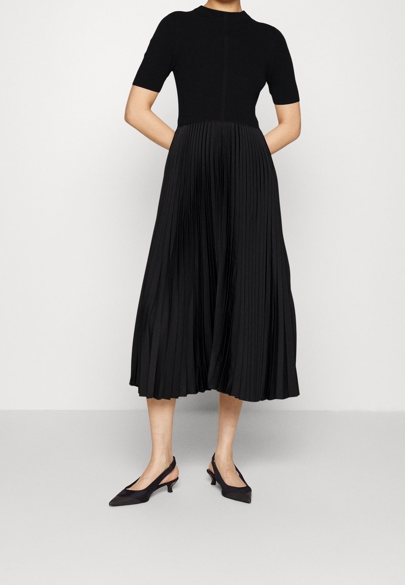 Femme portant un haut noir à manches courtes, une jupe midi plissée noire taille haute et des escarpins noirs à bride arrière à talons bas, debout devant un fond blanc.