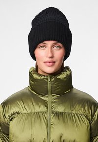 Gorro de malha preto canelado na cabeça de uma pessoa, combinado com um casaco acolchoado verde-oliva brilhante, com gola alta e fecho de correr.