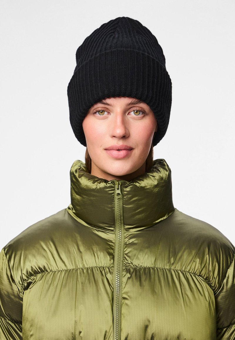 Gorro de malha preto canelado na cabeça de uma pessoa, combinado com um casaco acolchoado verde-oliva brilhante, com gola alta e fecho de correr.