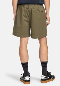 Olivgrüne Shorts mit elastischem Bund und einer Gesäßtasche. Getragen mit schwarzen Socken und Sneakers mit weißen und braunen Sohlen.