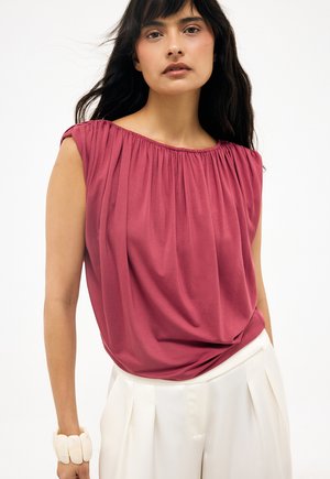 Žena s dlhými tmavými vlasmi nosí bezrukávový ružový nariasený top, biele plisované nohavice a masívny biely náramok na ľavom zápästí.