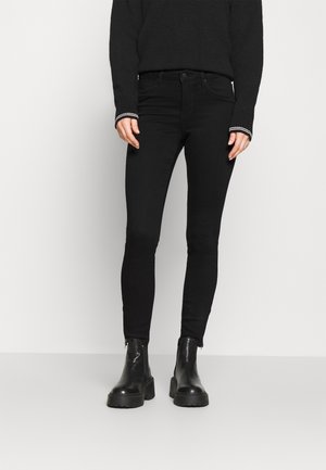 Osoba ubrana w czarne obcisłe jeansy, czarne masywne botki za kostkę oraz czarny sweter z mankietami w białe paski, stojąca przy białej ścianie.