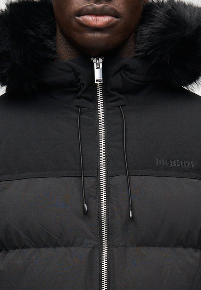 Casaco acolchoado preto com fecho de correr, com capuz destacável em pêlo sintético preto. Contrastes de textura notáveis entre as partes superior e inferior.