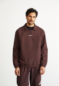 Vīrietis ar īsiem, cirtainiem matiem, ģērbies brūnā Adidas sporta uzvalkā ar logo uz krūtīm un biksēm, stāv pret baltu fonu.
