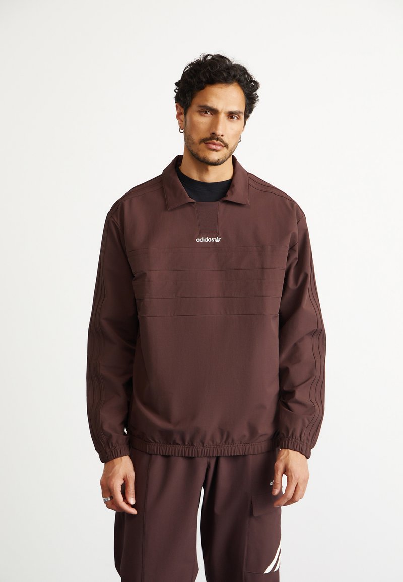 Vīrietis ar īsiem, cirtainiem matiem, ģērbies brūnā Adidas sporta uzvalkā ar logo uz krūtīm un biksēm, stāv pret baltu fonu.