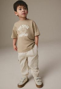 Tan bomulds t-shirt med hvid grafisk tekst, der siger "SJOVE TIDER HVER DAG," parret med lysebeige cargo bukser og grå sneakers med velcro remme.